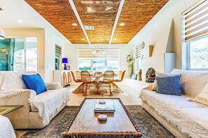 Uma sala de estar moderna com teto de madeira, dois sofás, uma mesa de centro e uma área de jantar com cadeiras de vime.