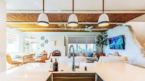 Une cuisine moderne dotée d’un îlot central, un coin repas avec des chaises en osier et un espace de vie comprenant un canapé et un téléviseur.