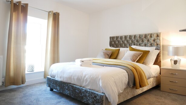 A neatly made bed with a tufted headboard, a bedside lamp, and a view of the window with curtains. - Gill Gardens Penthouse (Ulverston)