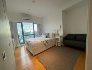 Enjoy a city view from the comfort of a spacious bed in this stylish room, featuring a sofa, bedside table, and large window.