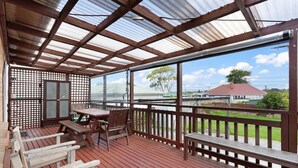 Un espace extérieur abrité, doté d’une terrasse en bois, d’une table et de chaises, et offrant une vue sur une maison située de l’autre côté d’une étendue d’eau.