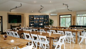 A spacious dining area with multiple wooden tables and white chairs, a large bookshelf filled with books, and a piano in the background. - Otter Creek Manor - Oversized 3bd, 1.5 Ba. 3 acres, creek access. (Eau Claire)