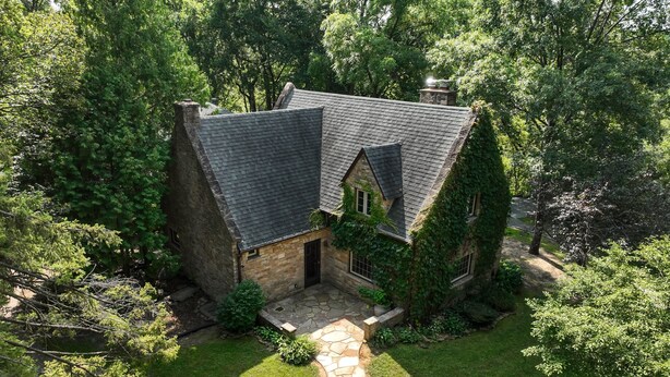 A stone house with a steeply pitched roof, surrounded by lush greenery and a stone pathway leading to the entrance. - Otter Creek Manor - Oversized 3bd, 1.5 Ba. 3 acres, creek access. (Eau Claire)