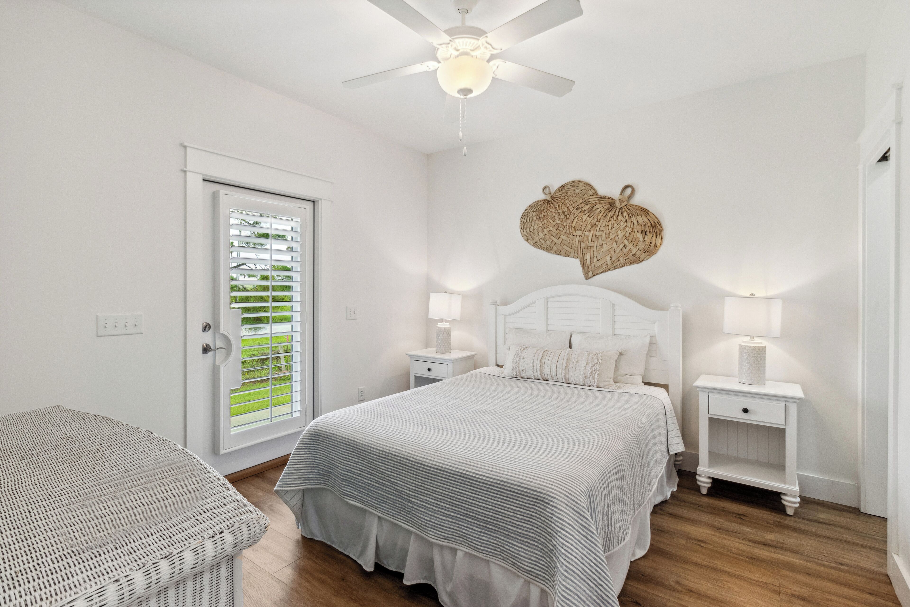 Une chambre à coucher comprenant un lit, deux tables de chevet, un ventilateur de plafond, une décoration murale en fibres tressées et une fenêtre avec des stores.