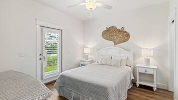 Une chambre à coucher comprenant un lit, deux tables de chevet, un ventilateur de plafond, une décoration murale en fibres tressées et une fenêtre avec des stores.