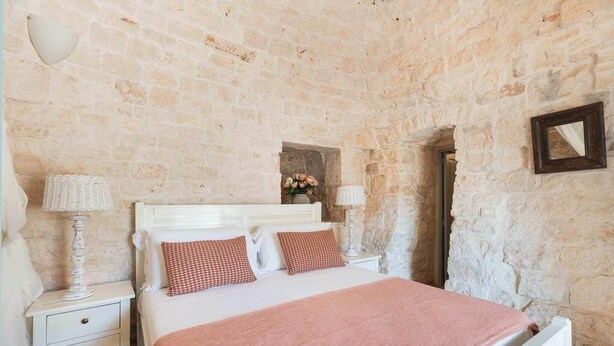 A bedroom with a stone wall, a white bed with checkered pillows, a bedside table with a lamp, and a mirror on the wall.