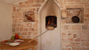 A stone fireplace with a pizza being baked inside, a wooden table with vegetables and bowls, and a hanging pan.