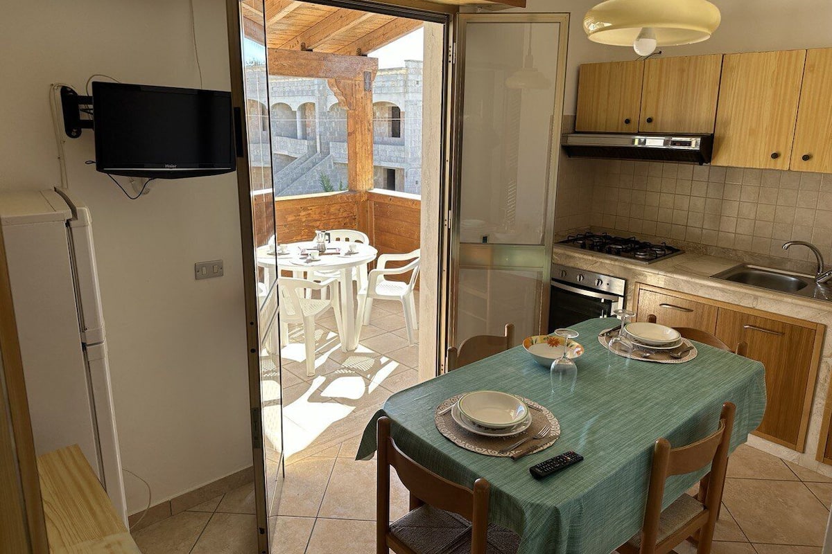 A compact kitchen with a dining area, a TV mounted on the wall, and a view of the outside balcony.