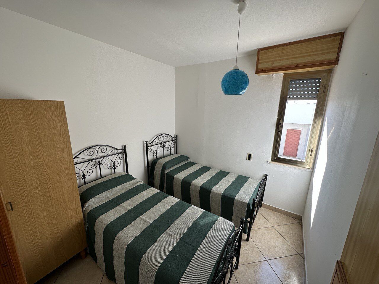 A small, clean room with two single beds, a wooden door, a window with blinds, and a hanging light fixture.