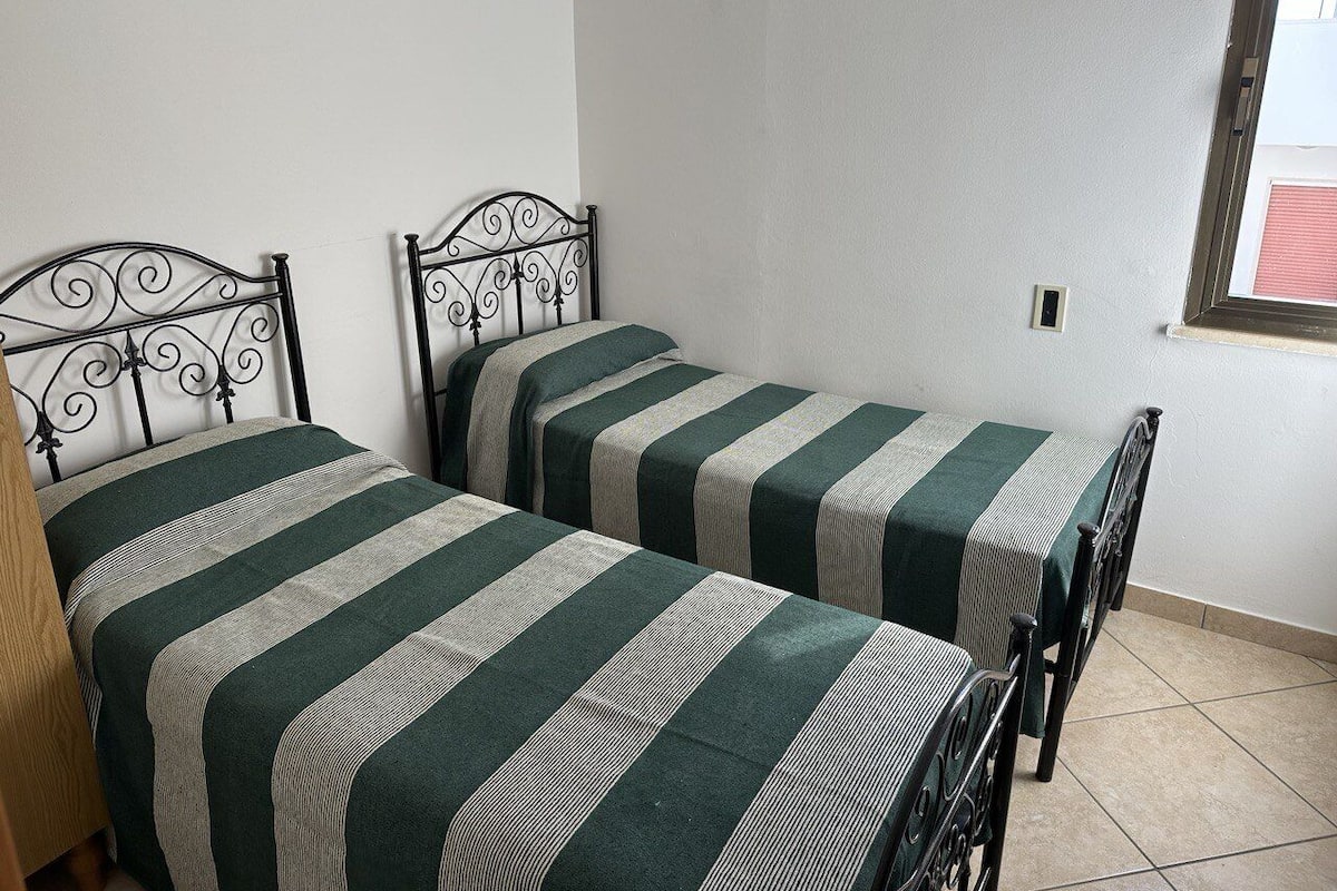Two single beds with metal frames and green striped bedding in a room with white walls and a tiled floor.