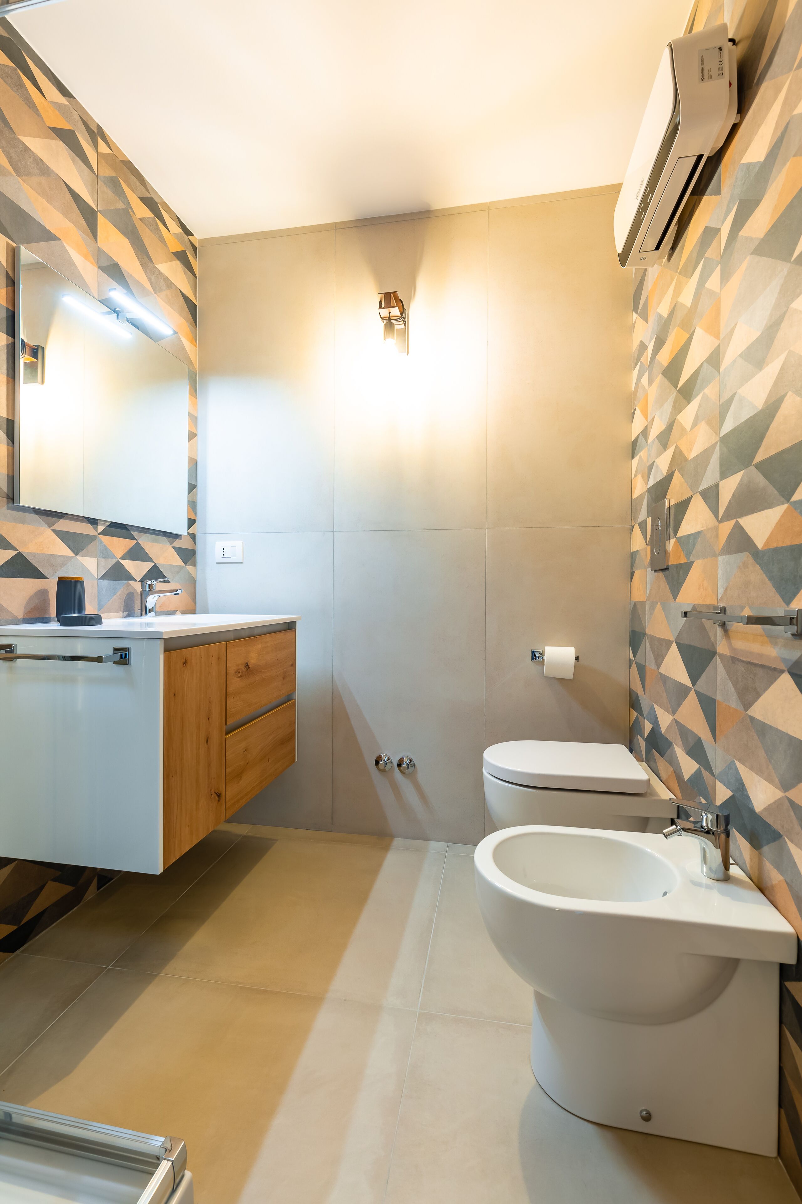 A modern bathroom with a white sink, wooden vanity, bidet, and a geometric-patterned wall.