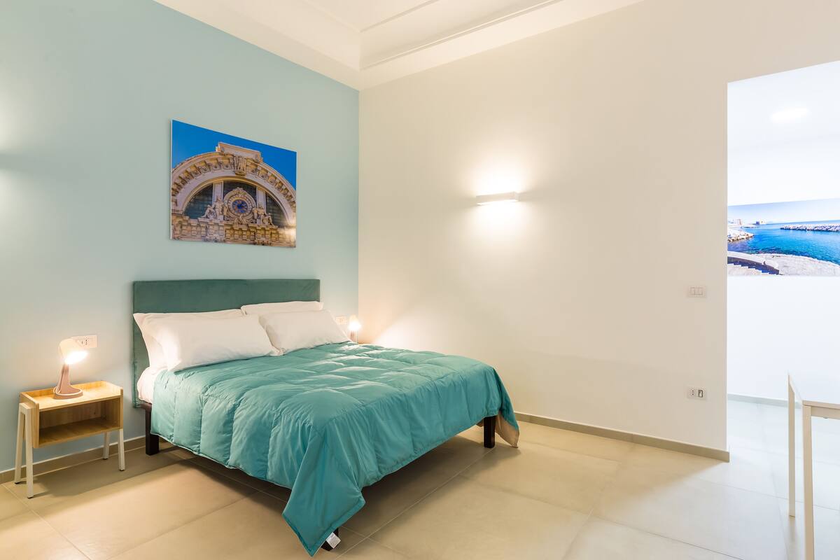 A bedroom with a bed, a nightstand, a wall-mounted lamp, and a painting of a coastal scene.