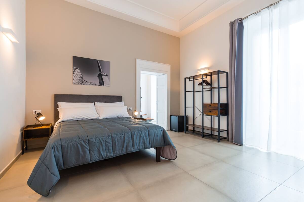 A modern bedroom with a large bed, a nightstand, a wall-mounted shelf, and a door leading to another room.