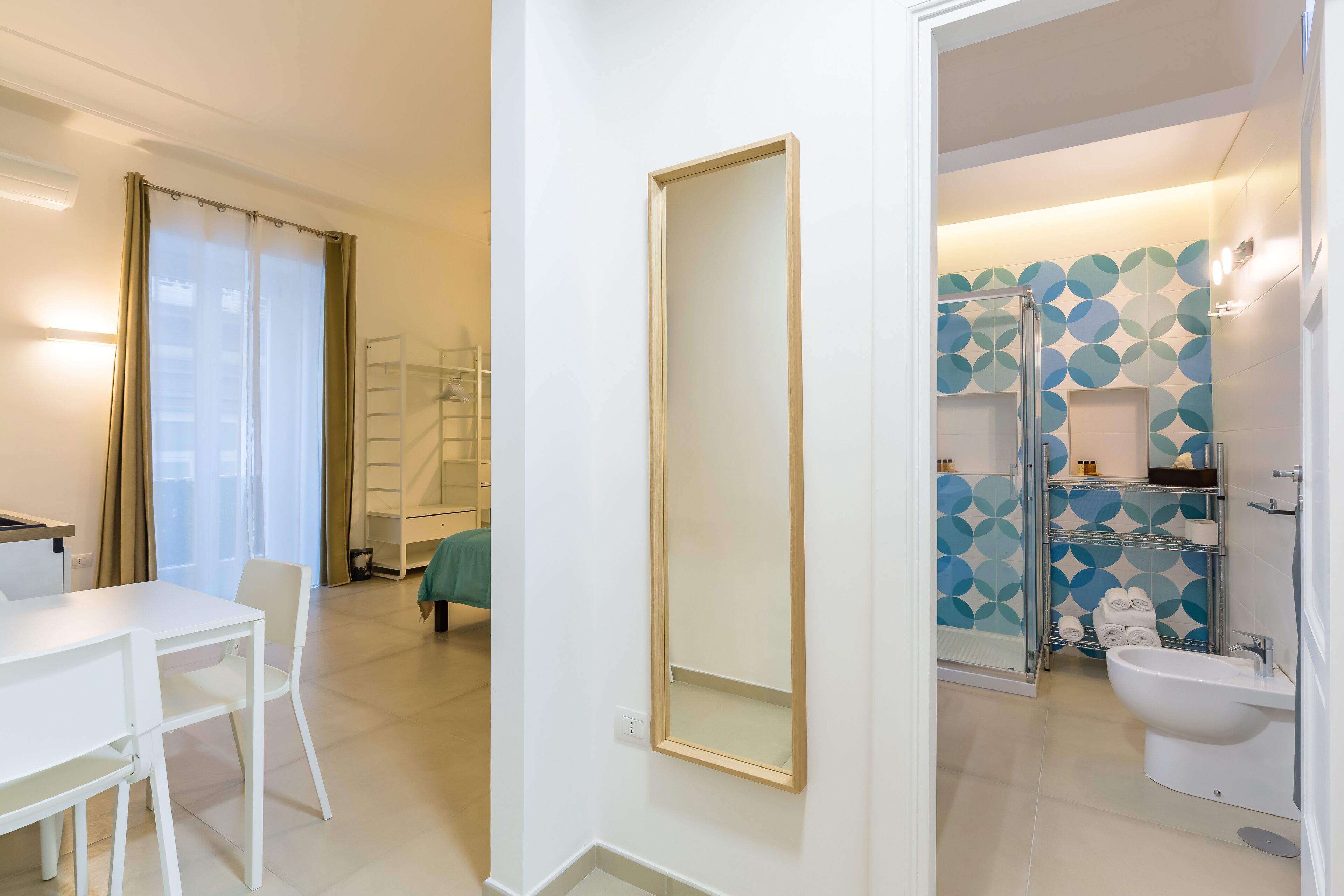 Una habitación de hotel moderna con zona de comedor, un espejo grande, una ducha con un salpicadero de azulejos geométricos y un lavamanos blanco.
