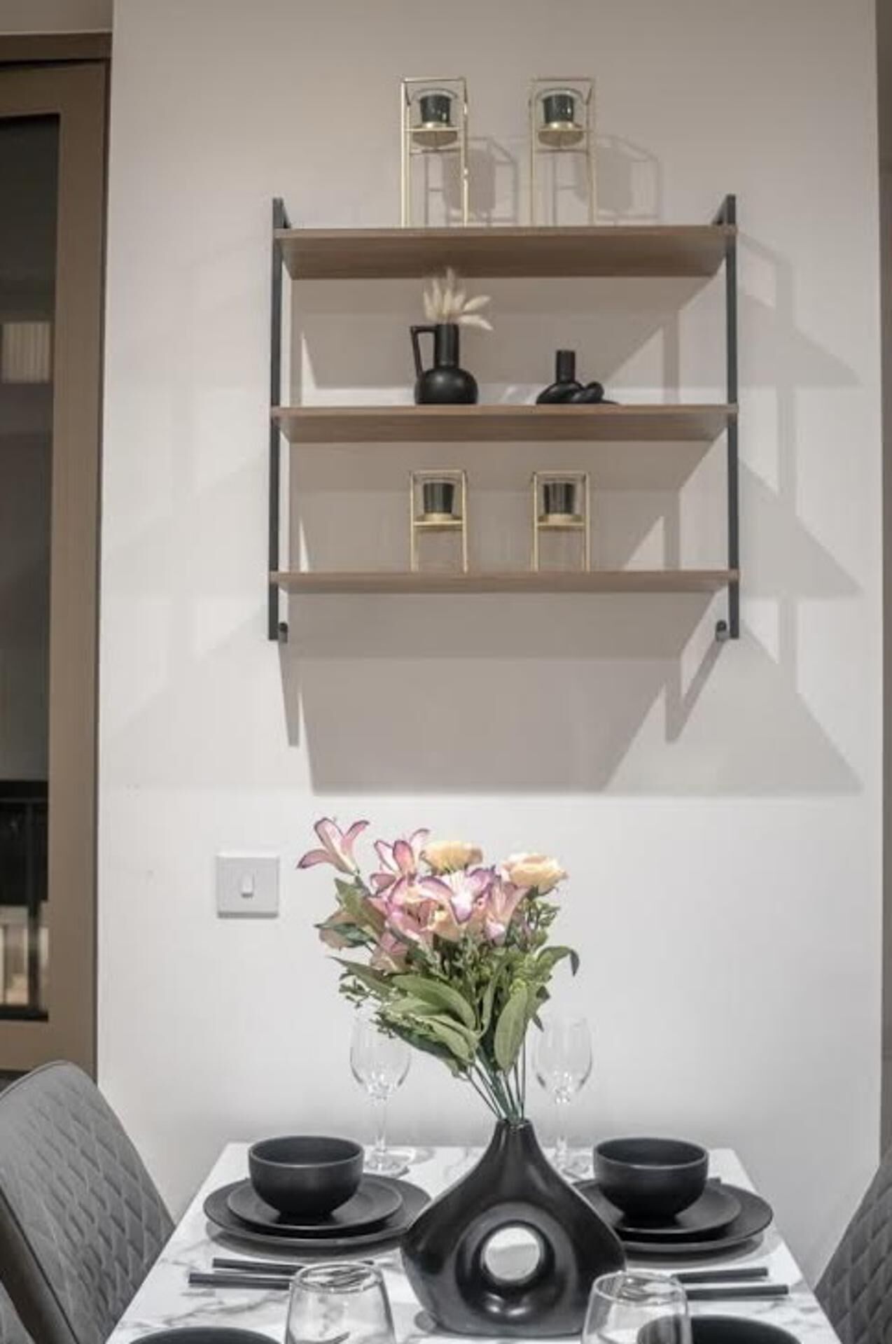 A dining area with a table set for two, a vase with flowers, and a wall-mounted shelf above.