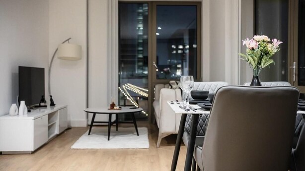 A modern living room with a dining area, a flat-screen TV, a vase with flowers, and a city view through large windows. - Immaculate 2-bed Apartment in Canary Wharf (London)