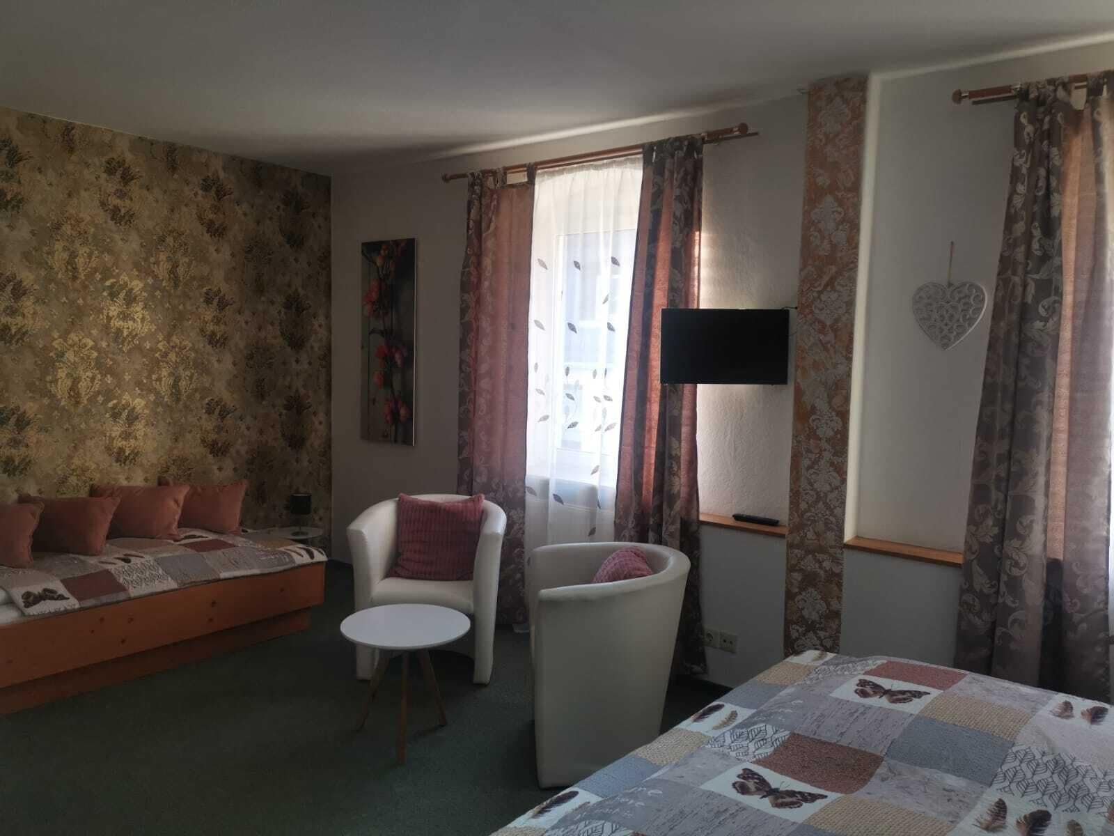 A cozy room with a bed, a sofa, a chair, and a small table. There is a painting on the wall and a TV mounted on the wall.