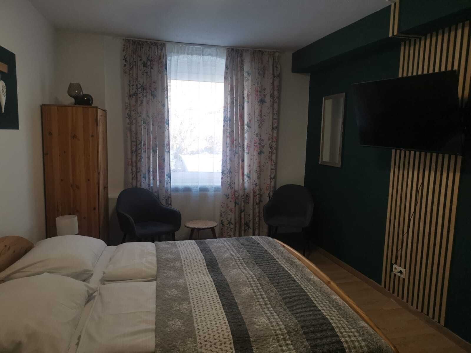 Quarto com uma cama, duas cadeiras, uma mesa pequena, uma televisão e um guarda-roupa.