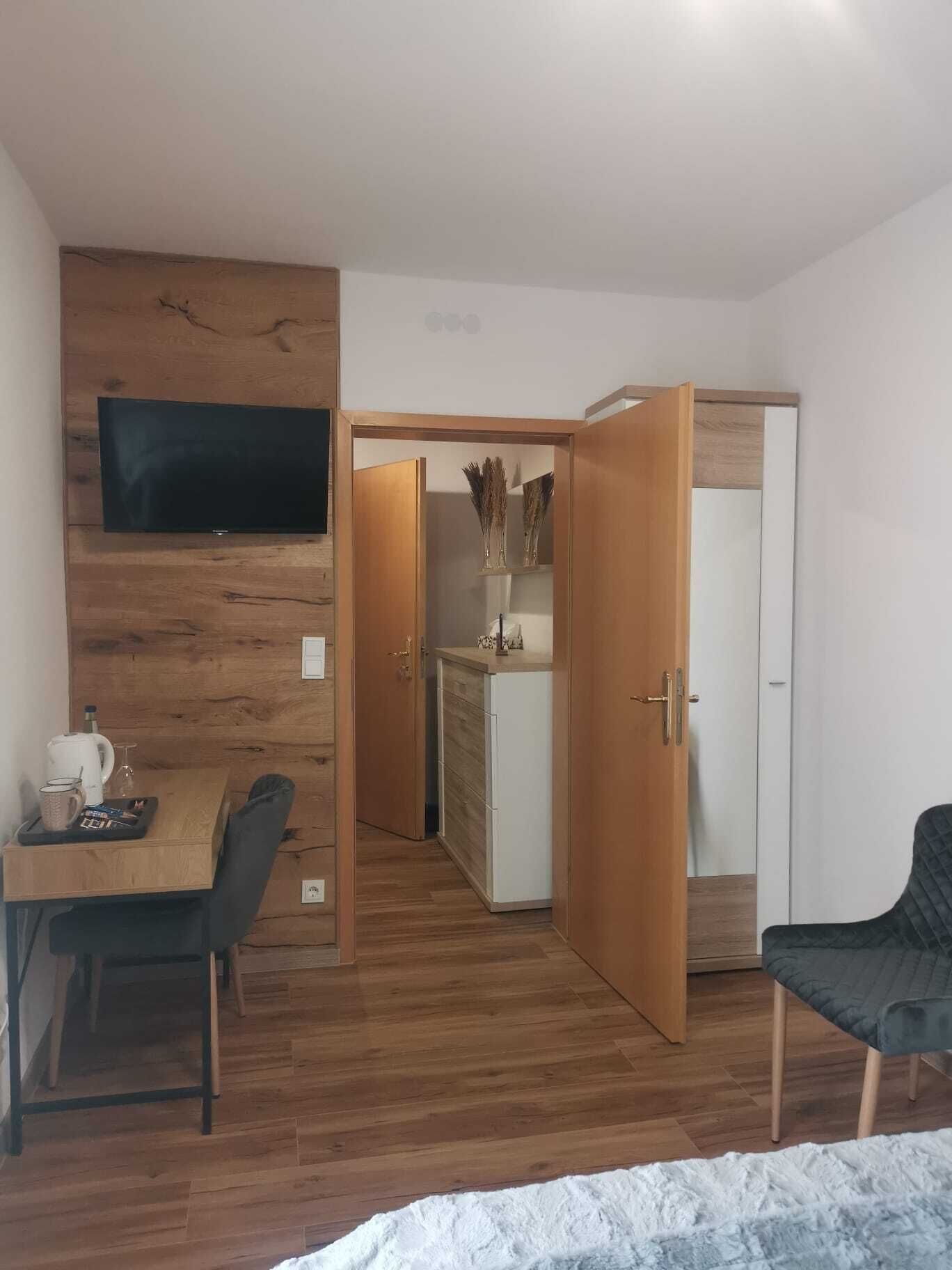 Una stanza moderna con parete in legno, TV a schermo piatto sopra un tavolo anch'esso in legno con una sedia, un mobile bianco e una porta che dà su un'altra stanza.