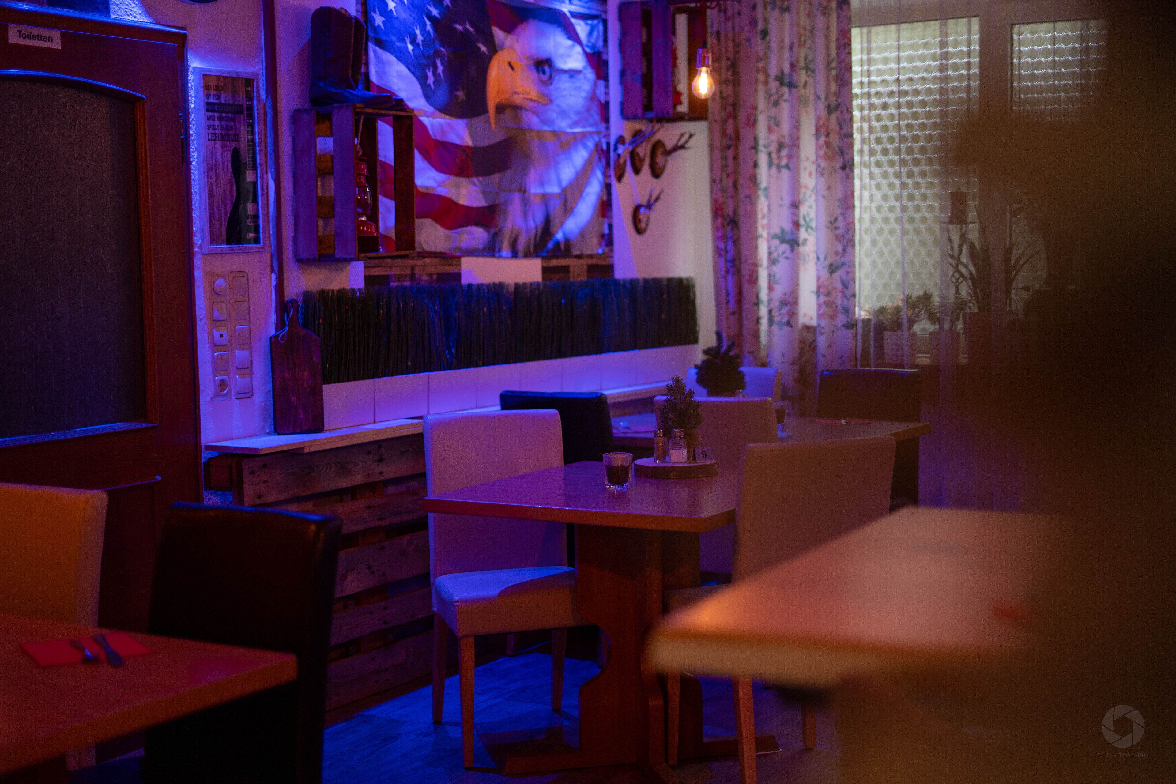 A dimly lit restaurant with wooden tables and chairs, a mural of an eagle, and a bar area with a counter and stools.