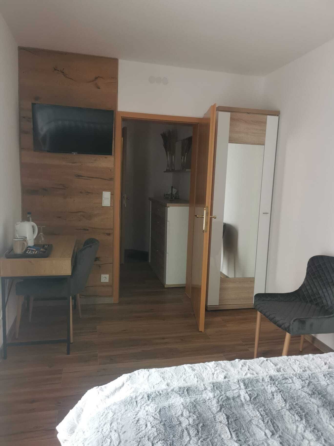 A hotel room with a wooden floor, a bed with a patterned blanket, a desk with a chair, a television mounted on the wall, and a wardrobe with a mirror.
