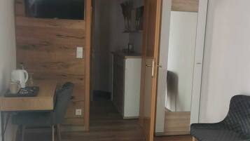 A hotel room with a wooden floor, a bed with a patterned blanket, a desk with a chair, a television mounted on the wall, and a wardrobe with a mirror.