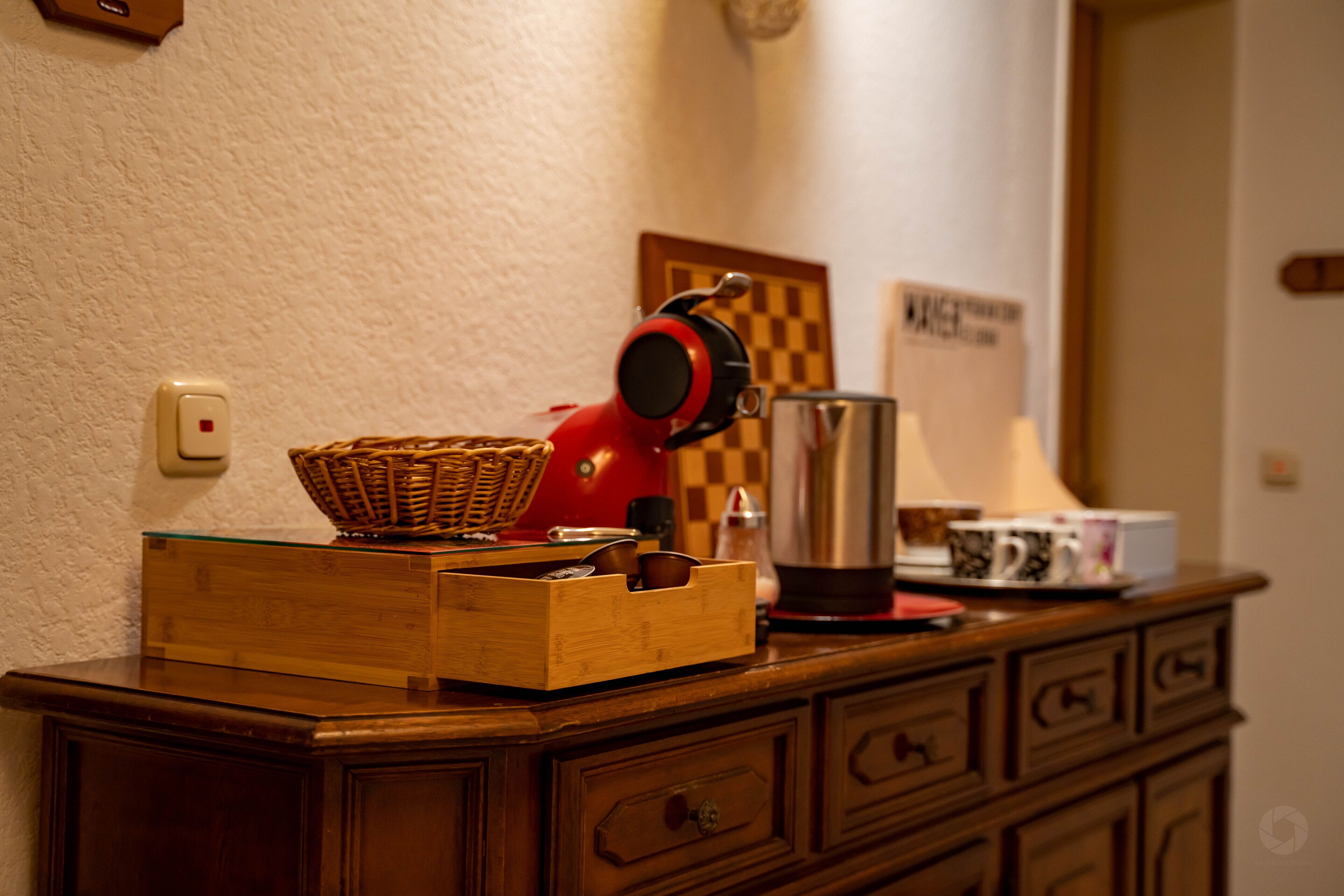 Een houten dressoir met lades, een rieten mand, een rode waterkoker, een roestvrijstalen koffiepot en een wanddecoratie met ruitjes.