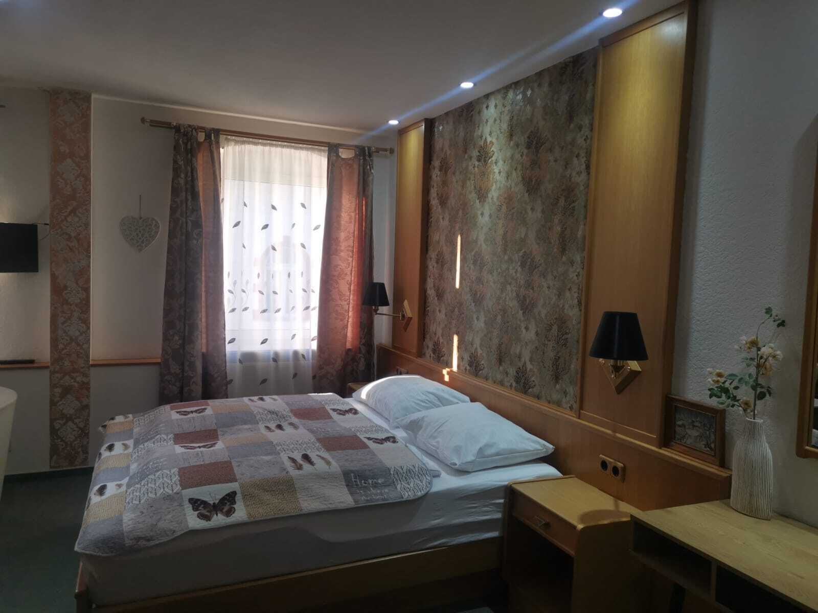 Une chambre d’hôtel avec un lit, une table de chevet, une commode, une lampe, un miroir et une fenêtre avec des rideaux.
