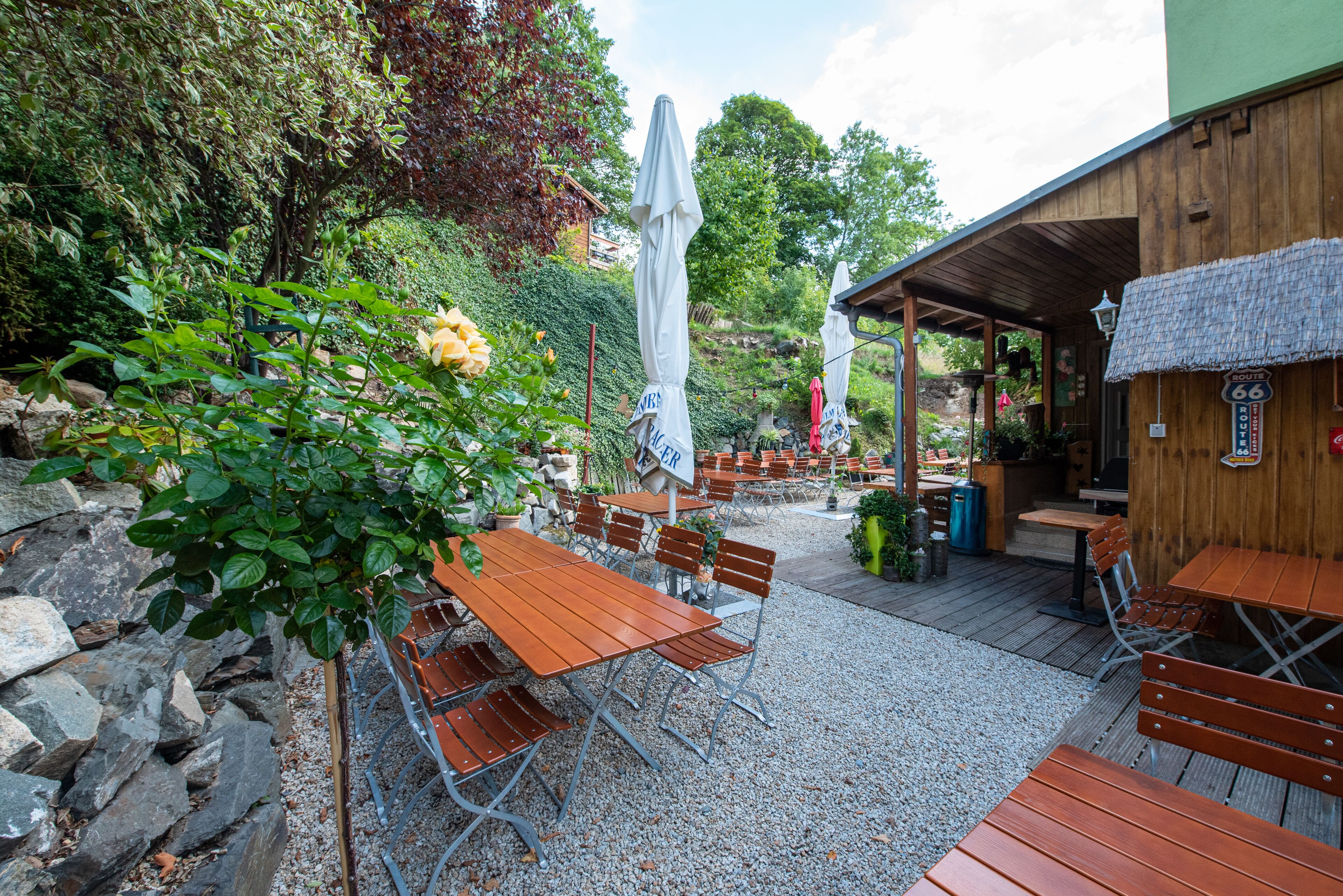 An outdoor patio area with wooden tables and chairs, umbrellas, and a rustic building with a sign.