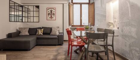Ein modernes Wohnzimmer mit einem Ecksofa, einem Esstisch mit roten Stühlen und einem großen Fenster mit Blick auf einen Kirchturm.