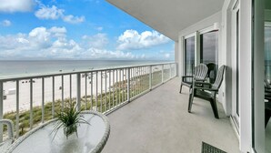 A balcony with a view of the beach and ocean, a round table with a plant, and two striped chairs.