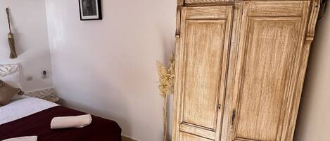 A room with a bed, a wardrobe, a wicker chair, and a vase with dried plants.