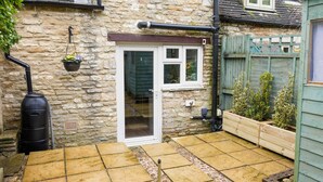 A stone house with a tiled patio, a wooden fence, and a hanging flower pot. - Cosy 1 bedroom cottage in The Cotswolds (Chipping Norton)