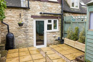 A stone house with a tiled patio, a wooden fence, and a hanging flower pot. - Cosy 1 bedroom cottage in The Cotswolds (Chipping Norton)