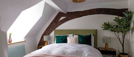 Un dormitorio con techo inclinado, vigas de madera a la vista, una cama con manta a rayas, dos lámparas de noche y una planta en maceta.