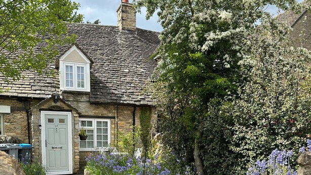 A stone cottage with a white door, surrounded by a garden with purple flowers and a tree with white blossoms. - Cosy 1 bedroom cottage in The Cotswolds (Chipping Norton)