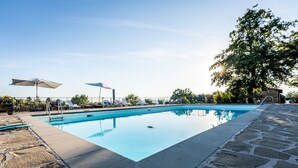 Uma piscina retangular com borda de pedra, rodeada por espreguiçadeiras e guarda-sóis.
