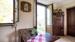 Uma mesa de madeira rústica com maçãs e um pano, um armário de madeira com objetos decorativos, e uma janela com vista para o verde.