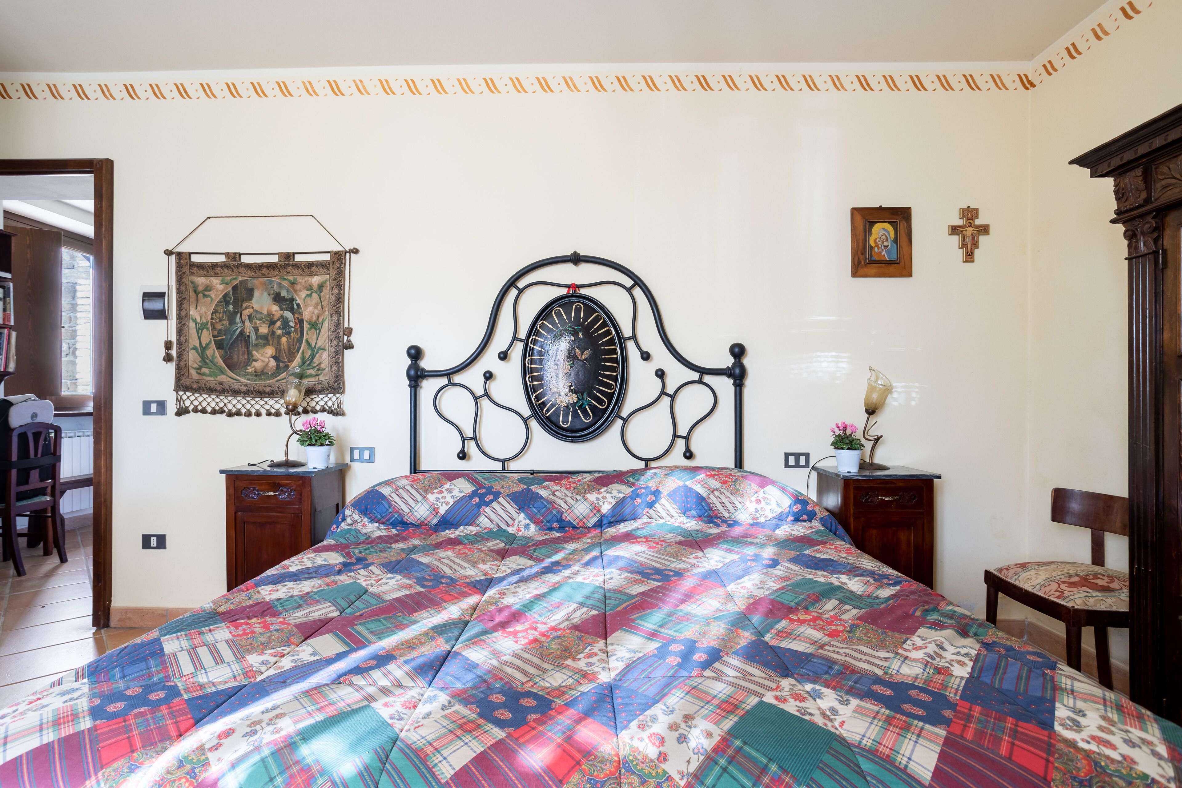 A bedroom with a large bed, a headboard with a mirror, a tapestry, a cross, and a small table with a lamp.