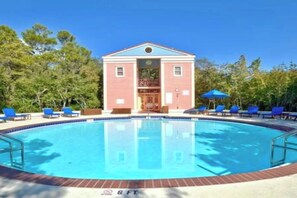 En swimmingpool med en byggnad i bakgrunden, omgiven av träd och blå solstolar.