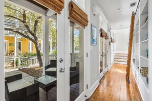 Un couloir doté d’un plancher en bois, de murs blancs et d’une porte en verre donnant sur un patio extérieur aménagé avec des meubles en osier et un arbre.