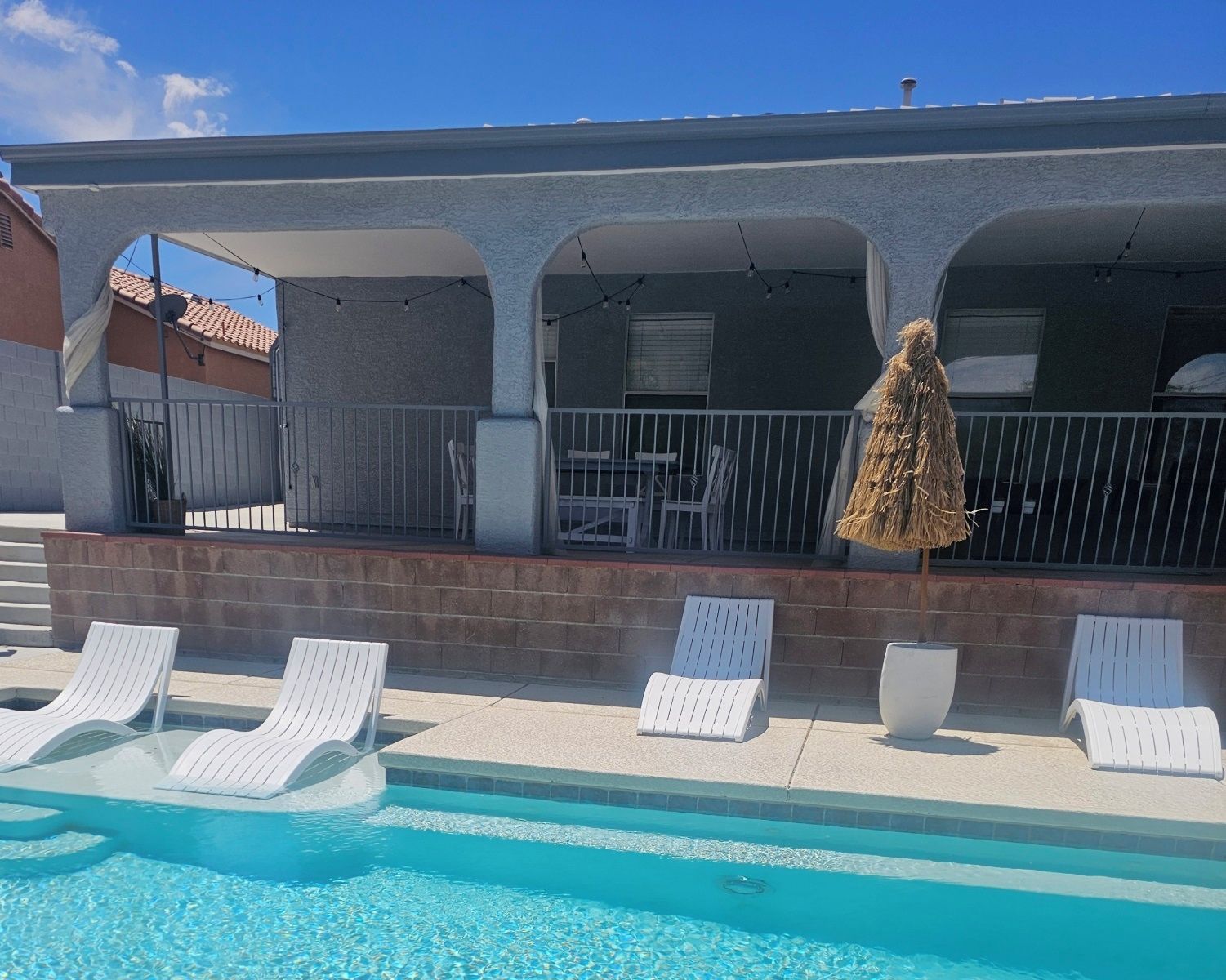 A pool area with white lounge chairs and a covered seating area with a straw umbrella.