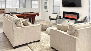A modern living room with a pool table, two sofas, a fireplace, and a ceiling fan.