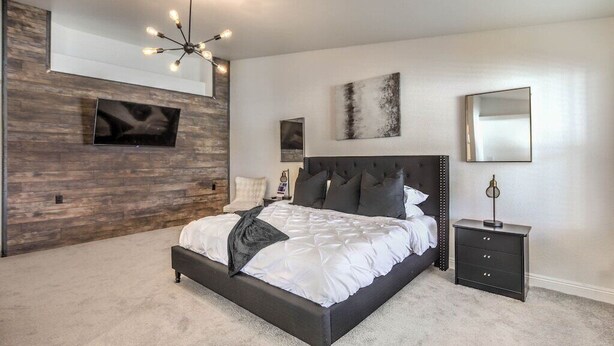 A modern bedroom with a large bed, a dark headboard, a mounted TV, and a wall-mounted light fixture.