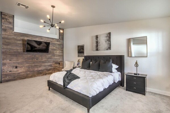 A modern bedroom with a large bed, a dark headboard, a mounted TV, and a wall-mounted light fixture.