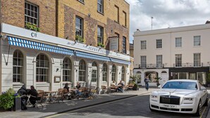 Un immeuble à la façade en briques, un café avec des tables en terrasse et une voiture blanche garée devant.
