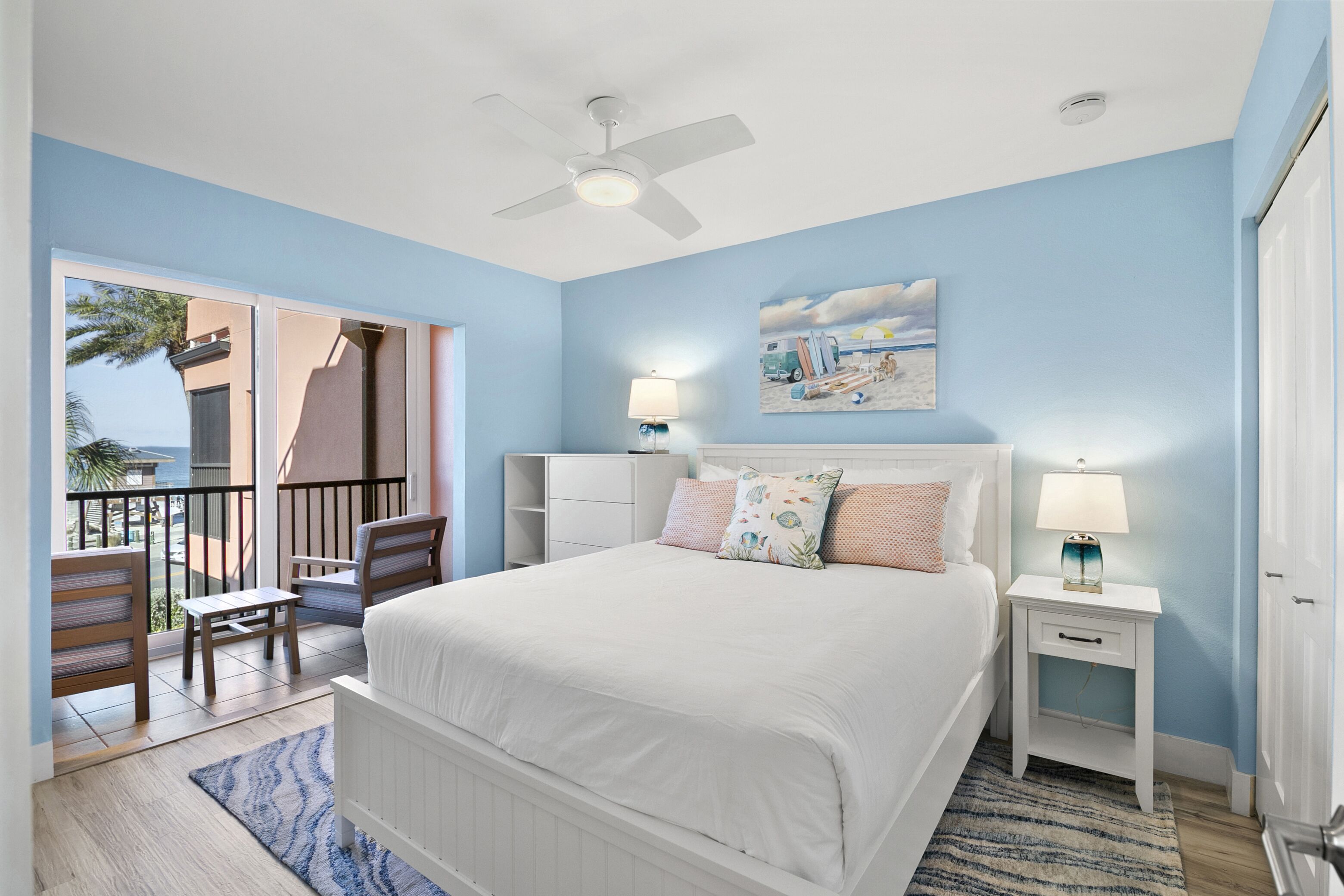 A bedroom with a bed, nightstands, a ceiling fan, a painting, and a view of the outdoors.