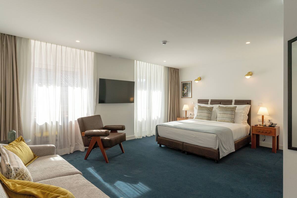 Una habitación de hotel con cama, sofá, sillón, televisión y mesita de noche con lámpara.