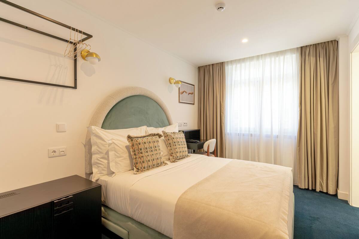Una habitación de hotel con cama, escritorio, silla, televisión y ventana con cortinas.