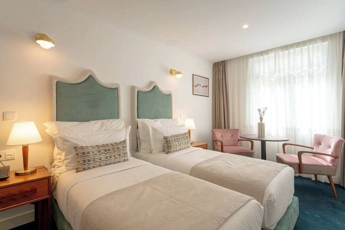 Habitación de hotel con dos camas, una mesita, dos sillas y un ventanal con cortinas.